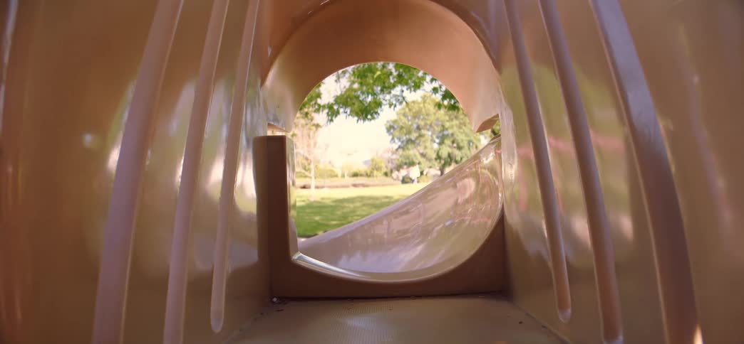 A happy little girl in a pink dress looks back at the camera before sliding down a tunnel slide at the park