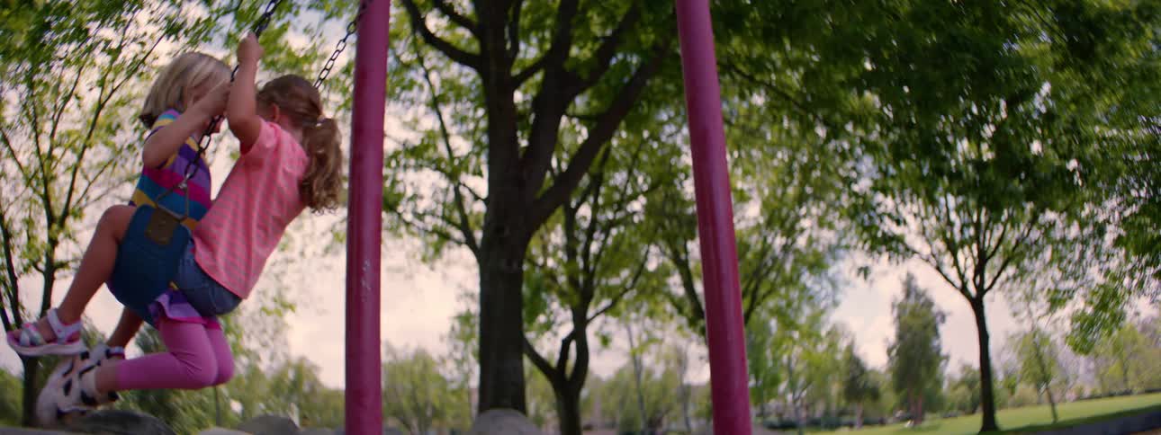 Two happy young girls swing together on a swing set at a park in slow motion