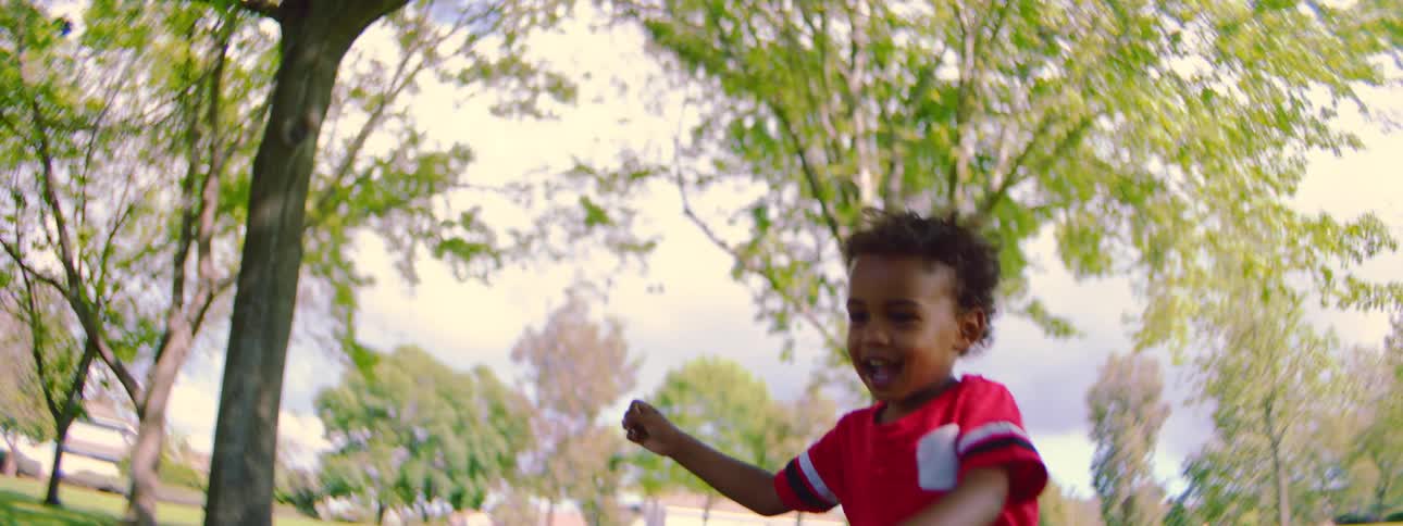 A joyous young boy with curly hair in a red shirt chases and plays with bubbles in a park in slow motion