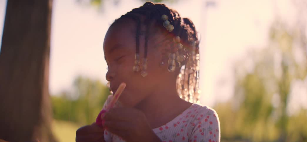 An adorable young girl with beaded braids eats a pink popsicle in a park during golden hour