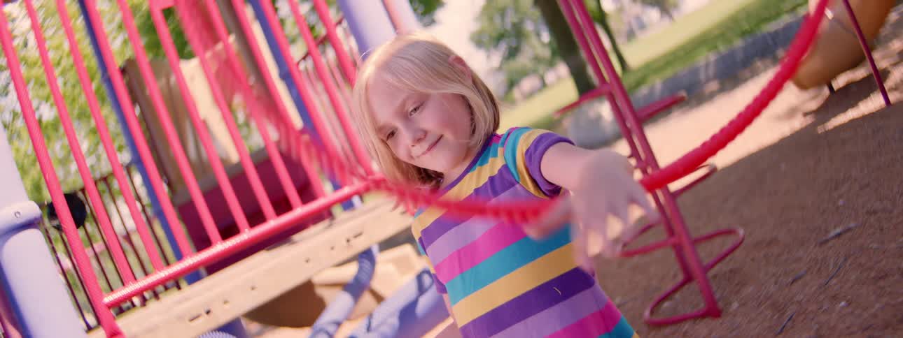 A happy young child in a colorful striped shirt smiles while spinning a red hula hoop in slow motion at a sunny playground
