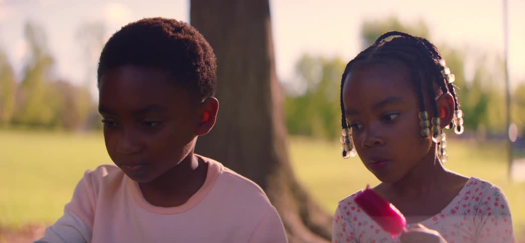 A close-up shot of a young boy and girl sitting together in a park while the girl eats a red popsicle
