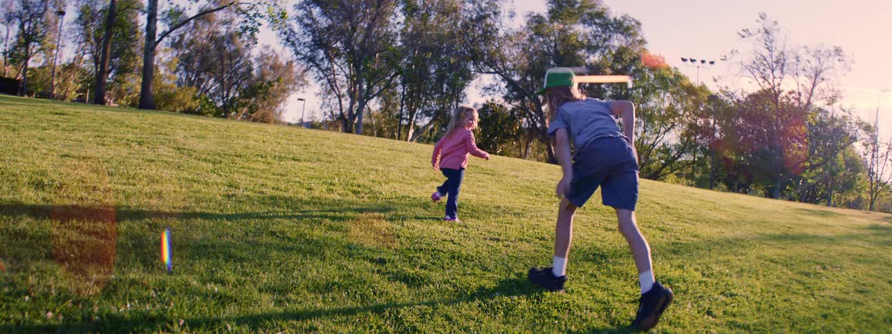A young boy and girl playfully race up a grassy hill in a sunlit park in slow motion