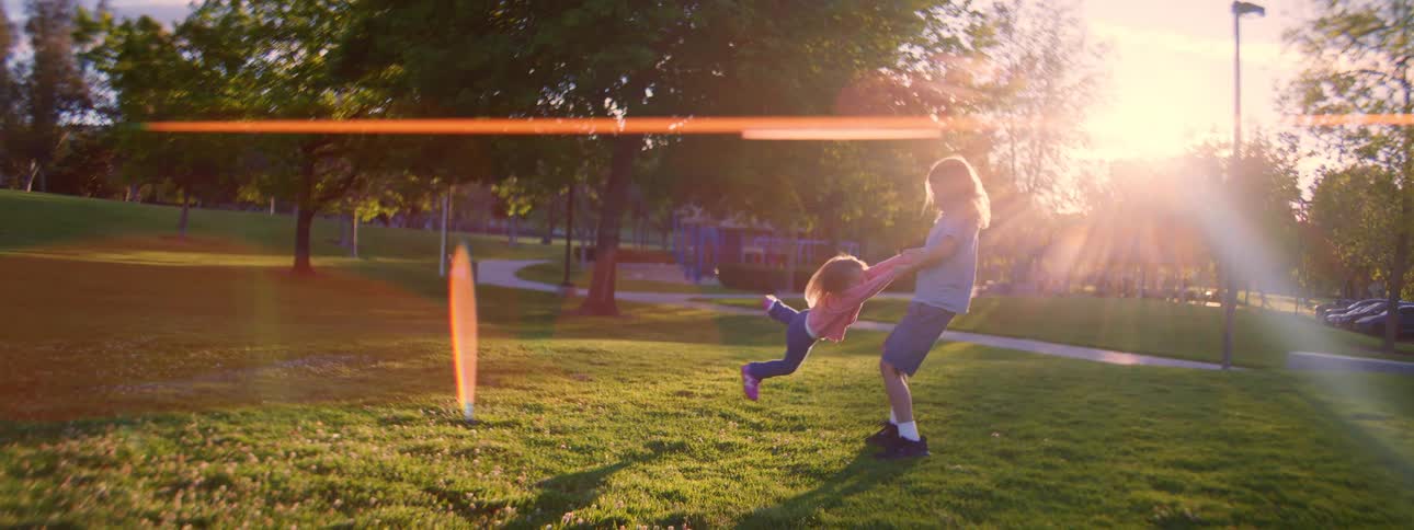 Carefree brother and sister spin in circles on a grassy lawn at a park as the sun sets, creating a beautiful lens flare