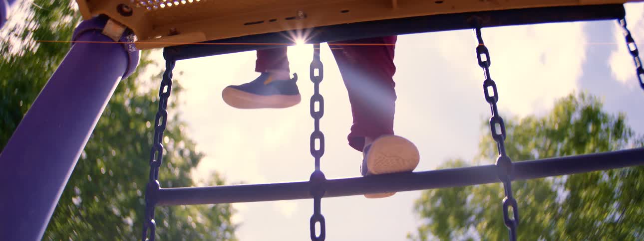 A slow-motion, low-angle shot captures a child's feet carefully climbing a playground ladder on a sunny day