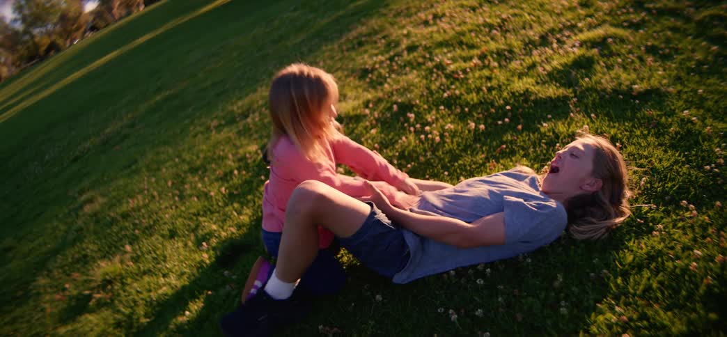A happy little girl runs in slow motion across a grassy park into her older brother's arms, where they fall and laugh together during golden hour
