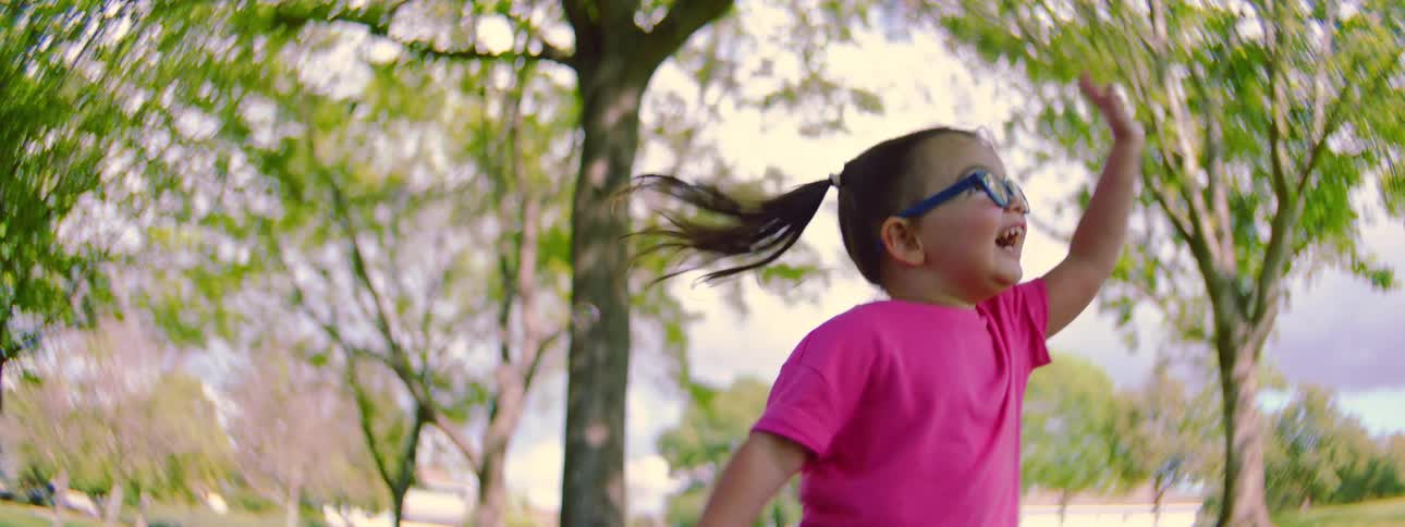A joyful toddler in a pink tutu and glasses happily plays with a bubble wand in a park in slow motion