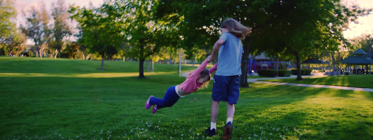 An older brother playfully spins his younger sister around in a sun-drenched park in slow motion