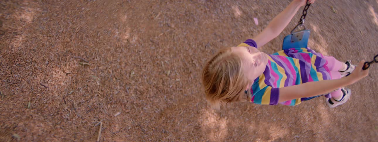 Slow motion overhead view of a young blonde girl looking up while gently swinging at the playground