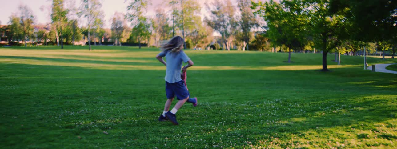 A young boy playfully spins his laughing little sister around on the lush green grass of a park in slow motion