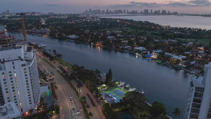 A breathtaking aerial timelapse captures the transition from dusk to night over a Miami waterway, with traffic flowing and the city skyline illuminating in the distance