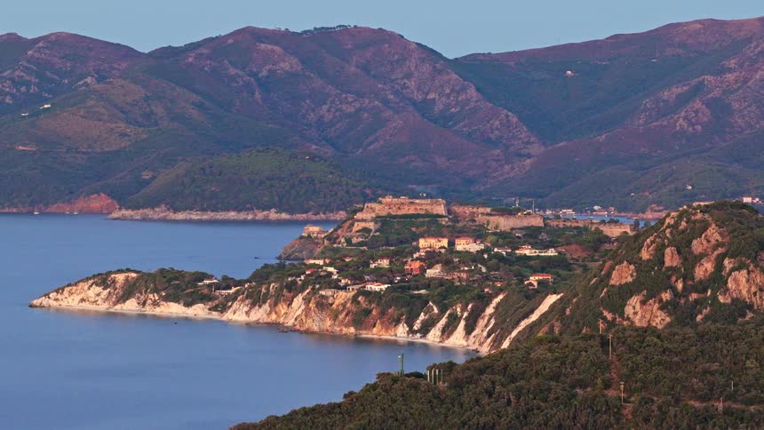 A sweeping aerial zoom-out of the historic coastal town of Portoferraio on the island of Elba, Italy, surrounded by mountains at dusk