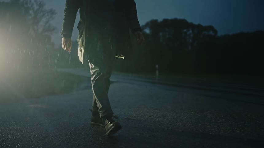 A person walks on a wet asphalt road at night in the rain, illuminated by car headlights in slow motion