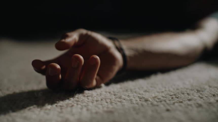 A dramatic close-up of a lifeless hand resting on the carpet in a dark room