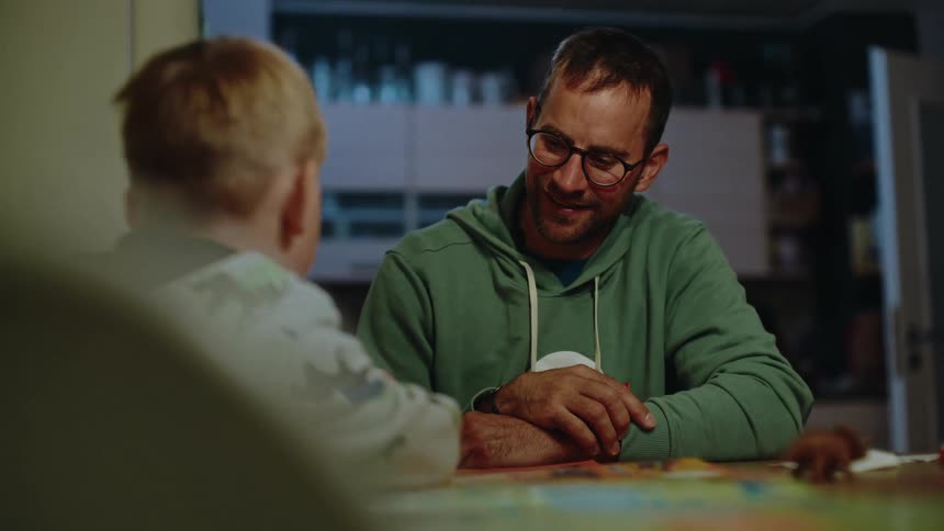 A loving father and his young son enjoy a quiet moment together, reading a book at the kitchen table in the evening