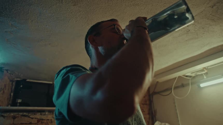 Thirsty craftsman in glasses hydrates from a water bottle in his rustic basement workshop, filmed in slow motion