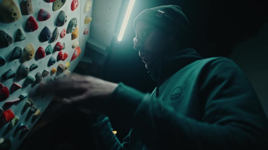 A male climber chalks up his hands in slow motion while studying a bouldering wall in a dark, dramatically lit gym