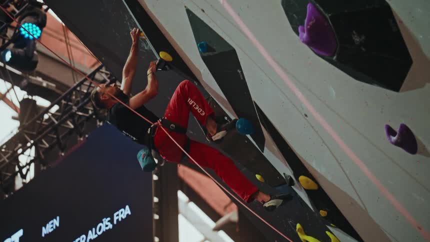 A professional male rock climber in red pants attempts a challenging route on an indoor competition wall before dramatically falling in slow motion
