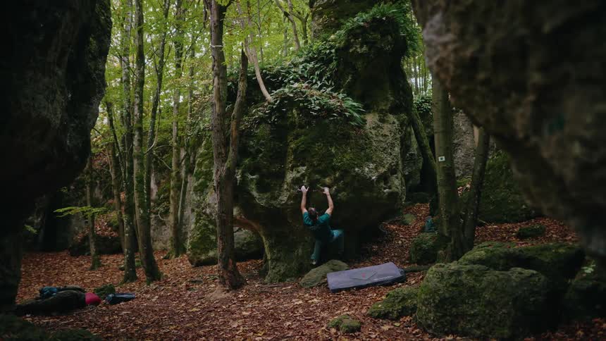 A slow tilt down through a lush green forest reveals a focused man bouldering on a large moss-covered rock