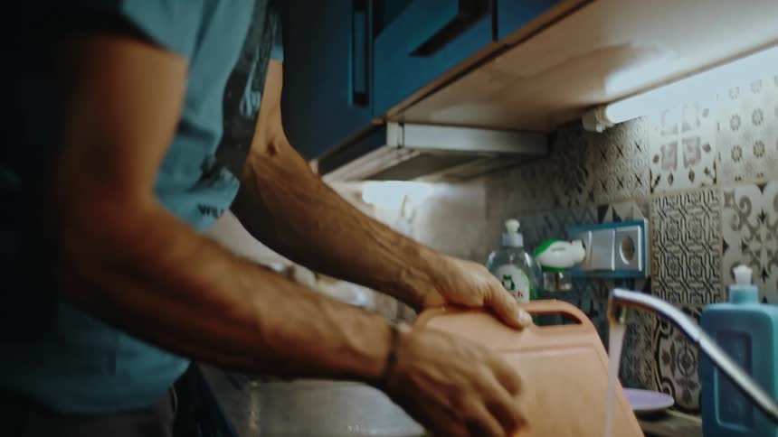 A man with glasses washes a cutting board with a sponge under running water in a dimly lit kitchen