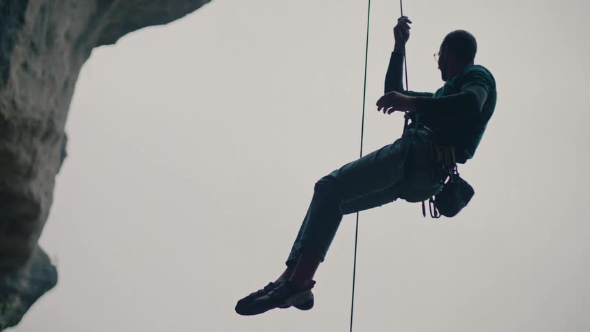 A silhouetted male rock climber carefully rappels down a large rock overhang as his female partner belays him from below