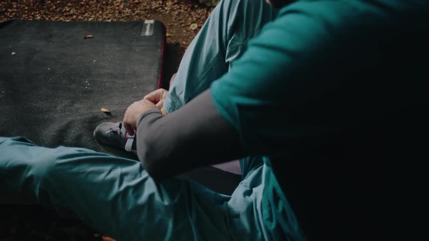 A male boulderer puts on his specialized climbing shoes in slow motion while sitting on a crash pad in the forest