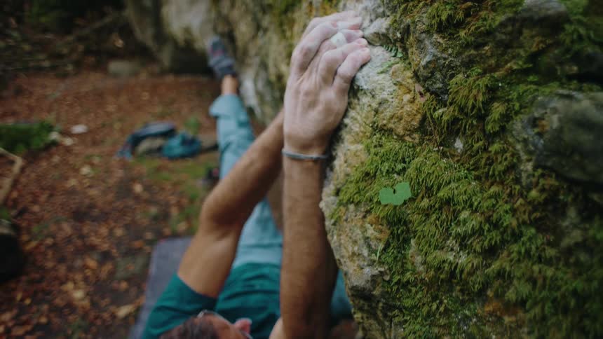A focused male climber tackles a challenging bouldering problem on a moss-covered rock in a serene forest setting
