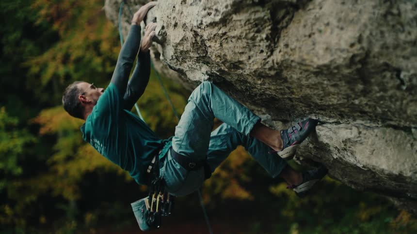 A determined rock climber in slow motion carefully navigates a difficult overhanging cliff face surrounded by autumn trees