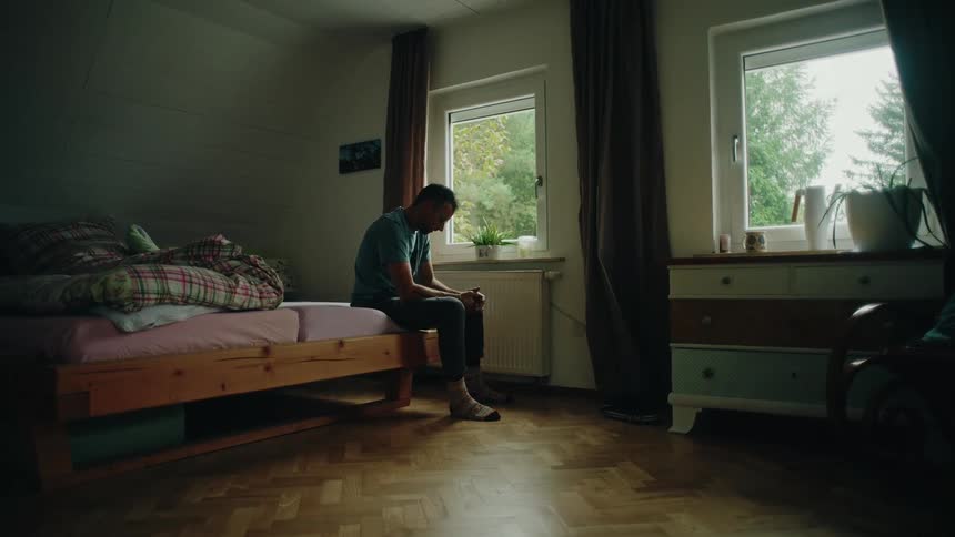 A depressed man sits alone on the edge of his unmade bed in a dark bedroom, staring out the window in silent contemplation