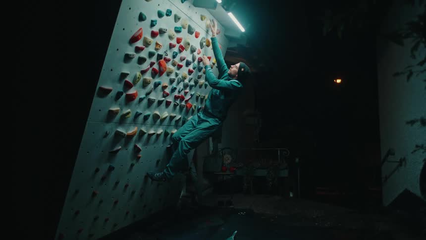 A man in a beanie slowly ascends a home bouldering wall during a focused nighttime training session