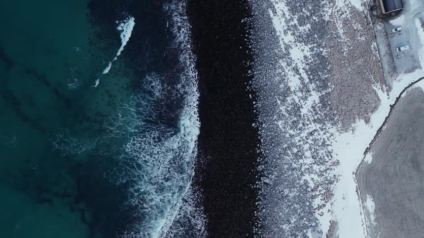 An aerial top-down view slowly rises over a dramatic winter coastline where turquoise ocean waves crash against a rocky, snow-dusted shore