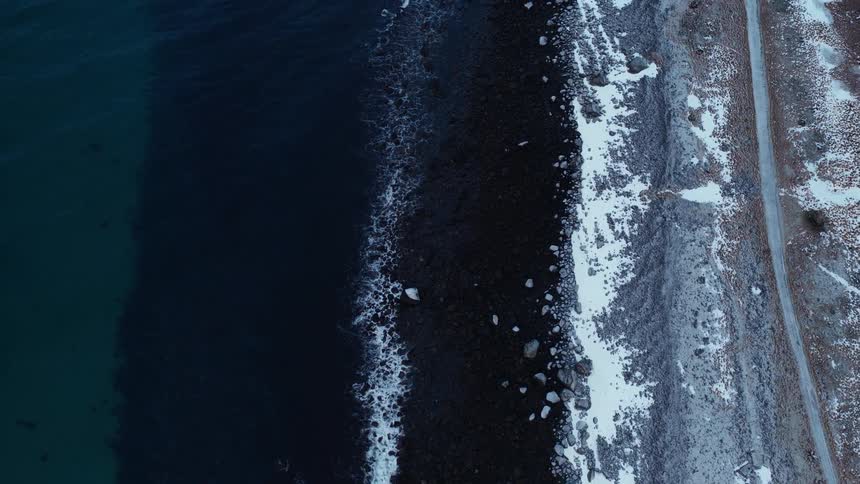 A top-down aerial view of a snowy, rocky coastline as dark blue ocean waves gently roll onto the shore