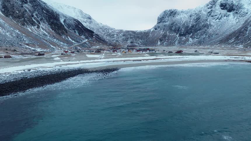 An aerial view reveals a quaint village on a snowy coastline, nestled between towering mountains and the cold, turquoise sea