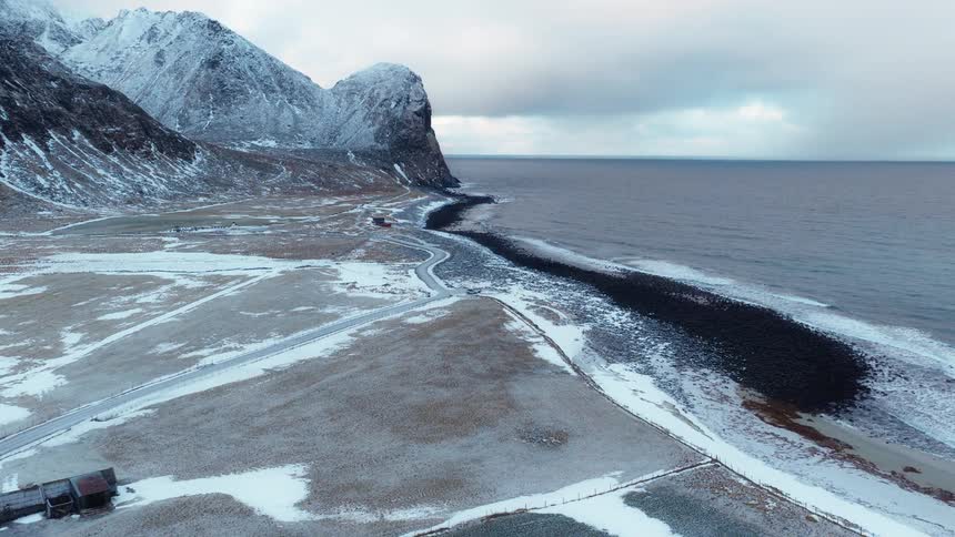 A dramatic aerial view of a remote coastal village in winter, where snow-covered mountains meet the vast, cold sea