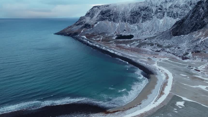 An epic aerial view pulls back to reveal a vast, snow-covered mountain range towering over a cold, teal-colored sea and a frozen shoreline