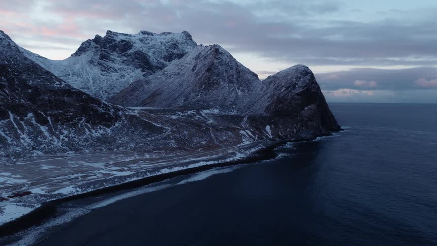 A majestic aerial view captures snow-covered mountains meeting the tranquil ocean along a remote coastline at dusk