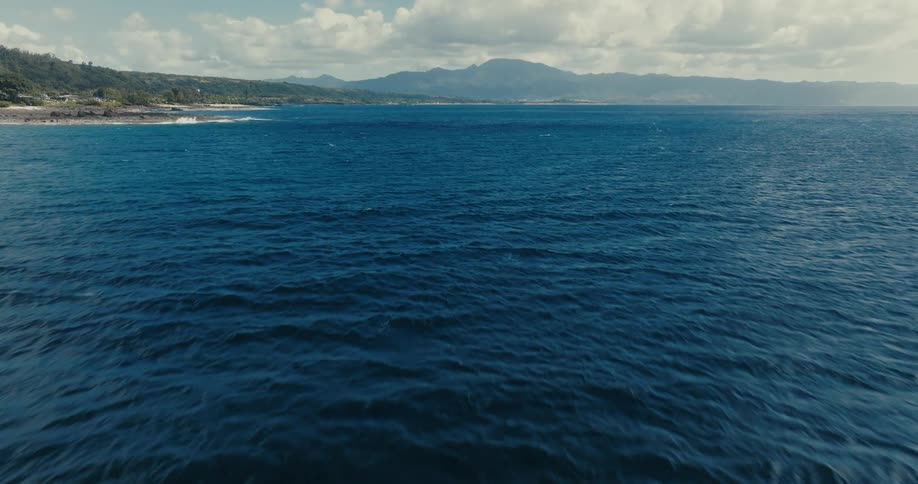 A fast aerial perspective glides low over the deep blue ocean waters towards a distant mountainous coastline