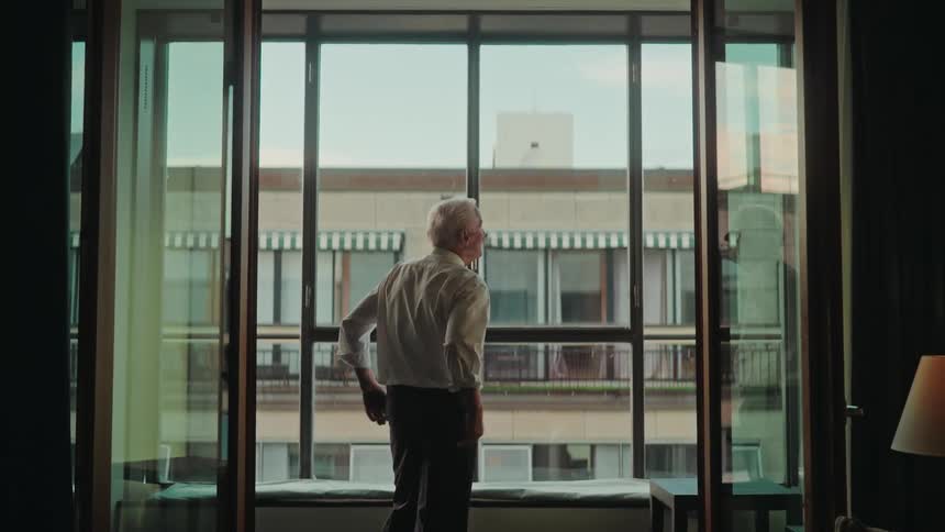 An elderly man stands contemplatively at a large window in his apartment, looking out at the city view with his hands behind his back