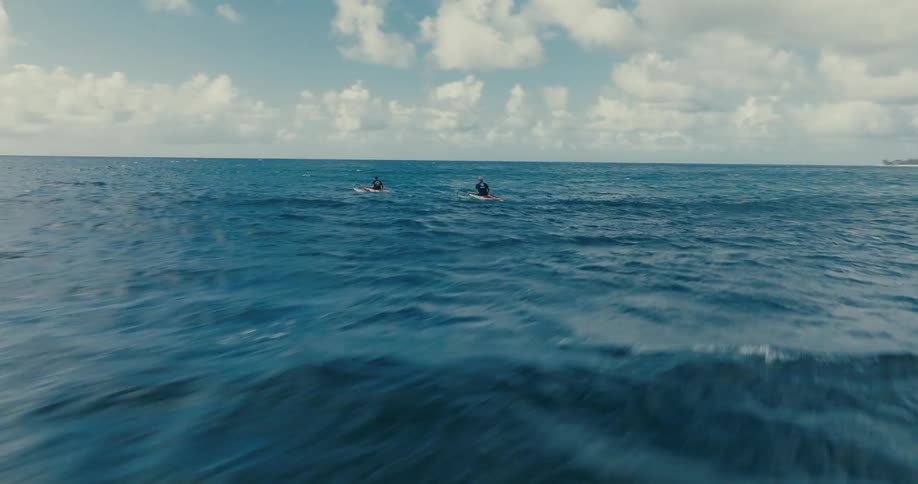 A dynamic low aerial shot flies over the water, following two people paddling kayaks in the open ocean near a tropical shore