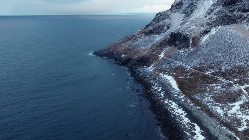A serene aerial view shows a snow-dusted rocky coastline meeting the vast, dark ocean under an overcast sky