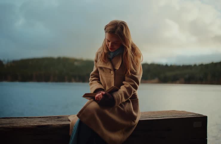 A serene young woman in a warm coat enjoys reading a book while sitting on a wooden dock by a calm lake on an overcast day