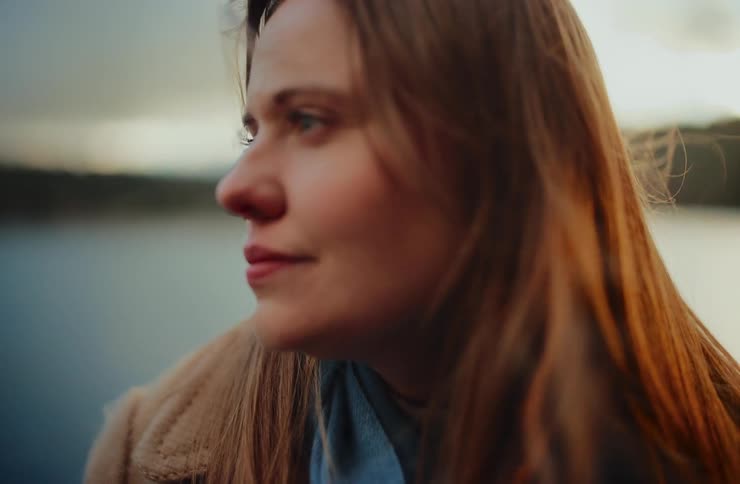 A pensive woman's face in profile is illuminated by the golden sunset light as wind blows through her hair by a serene lake