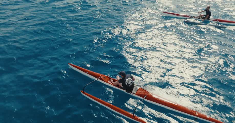 An exhilarating aerial view follows two men racing their outrigger canoes across the vast, sun-dappled open ocean