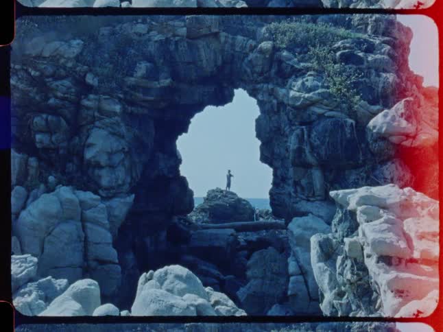 A lone figure stands on a rock framed by a natural sea arch in a vintage film clip that ends with a dramatic film burn