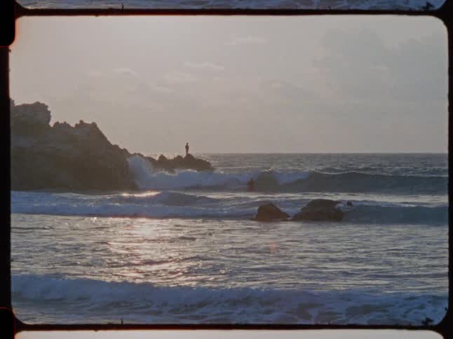 Vintage film captures a lone fisherman silhouetted on a rocky shore as waves roll in at sunset