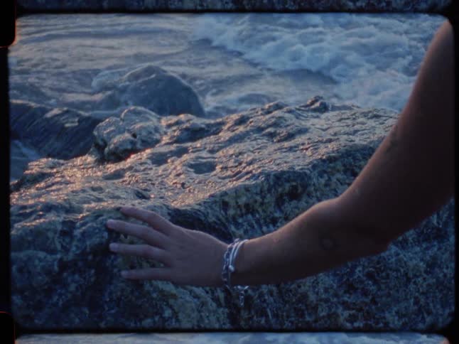A woman with blonde hair gently touches a sun-kissed rock on a rugged coastline, watching the waves at sunset