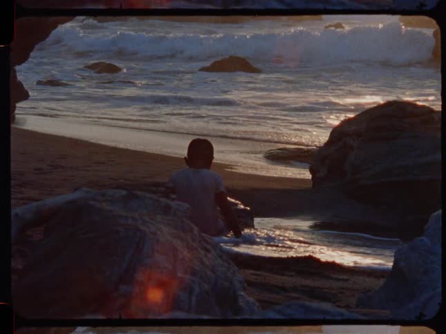Vintage footage captures a solitary boy on a rocky beach watching the gentle ocean waves during a beautiful sunset