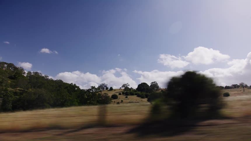 A high-speed tracking shot from a vehicle window captures the blurred, sunny landscape of rolling hills and dry fields