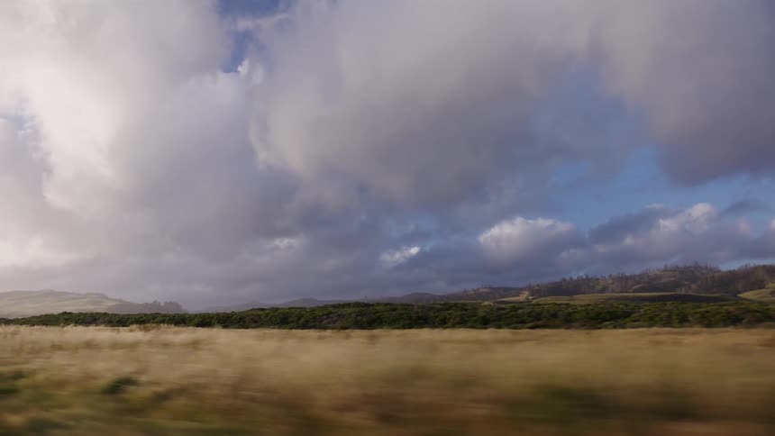 A misty coastal landscape with rolling green hills passes by as seen from a moving car under a dramatic cloudy sky