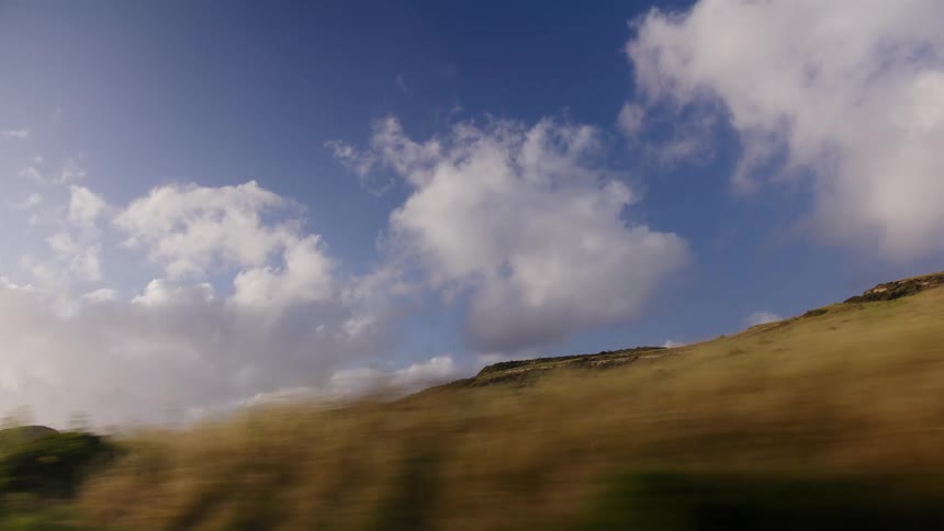A scenic point-of-view shot from a moving car captures the blur of a grassy roadside against a backdrop of rolling green hills and a bright blue sky